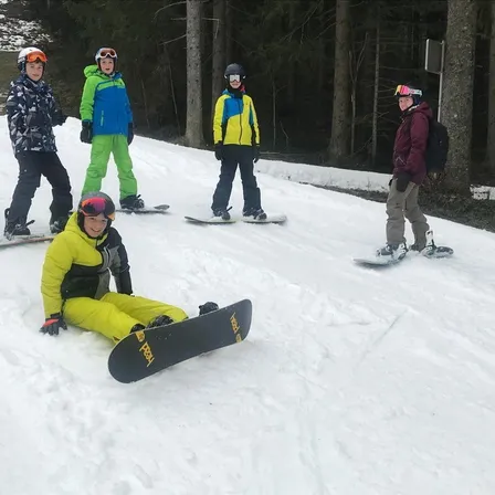 Vier Snowboarder stehen auf einem verschneiten Hang, einer in einem gelben Mantel sitzt, während die anderen mit Helmen und Handschuhen stehen.