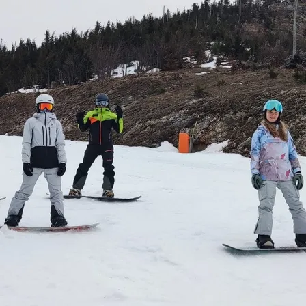 Eine Gruppe von Menschen steht auf Snowboards auf einem verschneiten Hang. Einige tragen Helme und Schutzbrillen. Der Boden ist felsiger Natur.