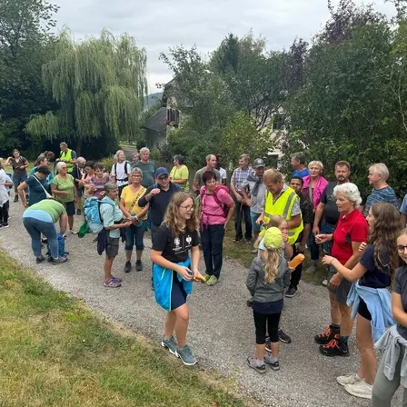 Eine Gruppe von Menschen steht auf einer Straße, vielleicht für eine Wanderung. Einige tragen Brillen und Turnschuhe, und andere haben Rucksäcke dabei. Eine Person hält ein gelbes Objekt. Ein goldenes Kreuz steht rechts.
