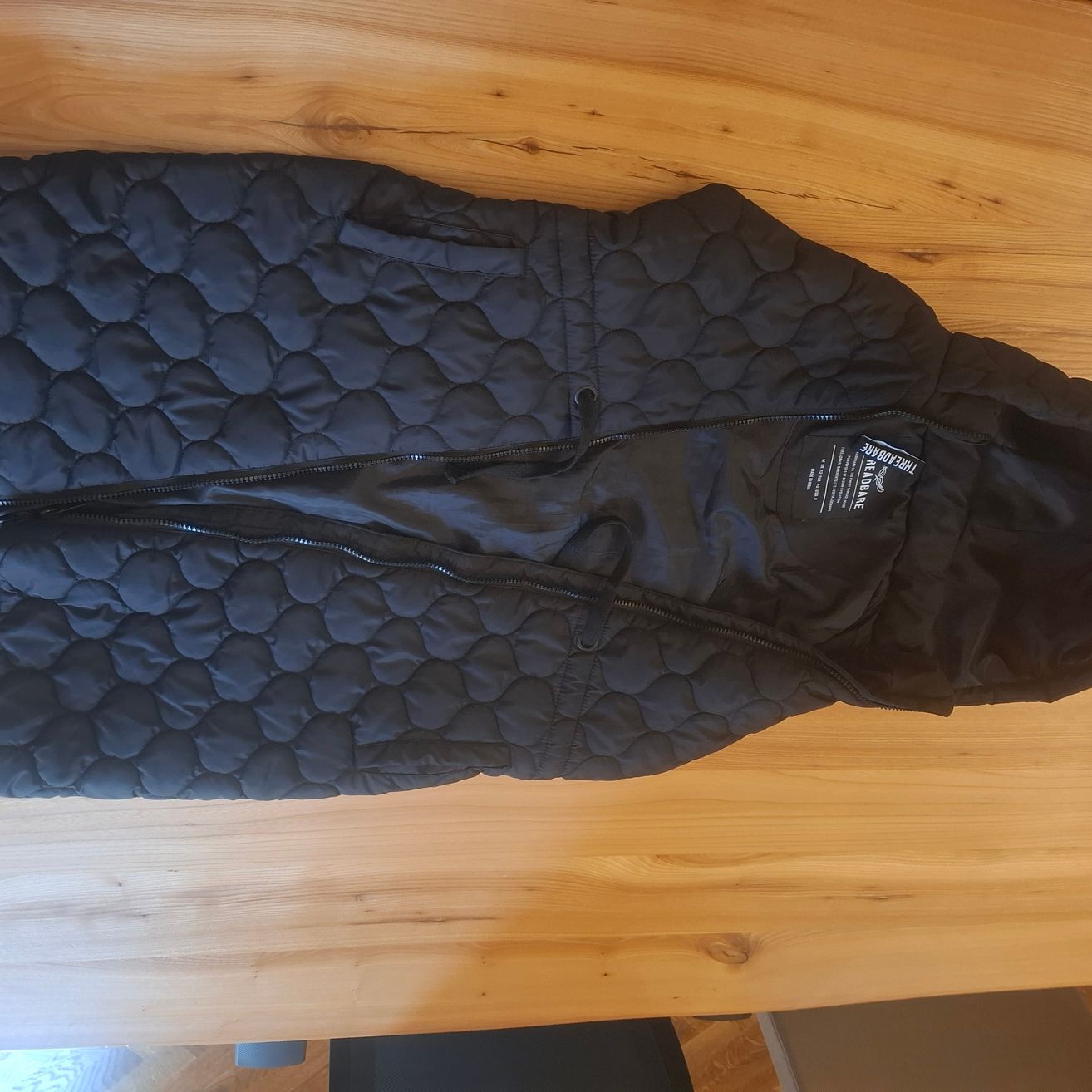 Eine schwarze, gesteppte Jacke mit zwei Reißverschlüssen liegt auf einem Holztisch. Die Jacke hat ein Logo auf der linken Seite.