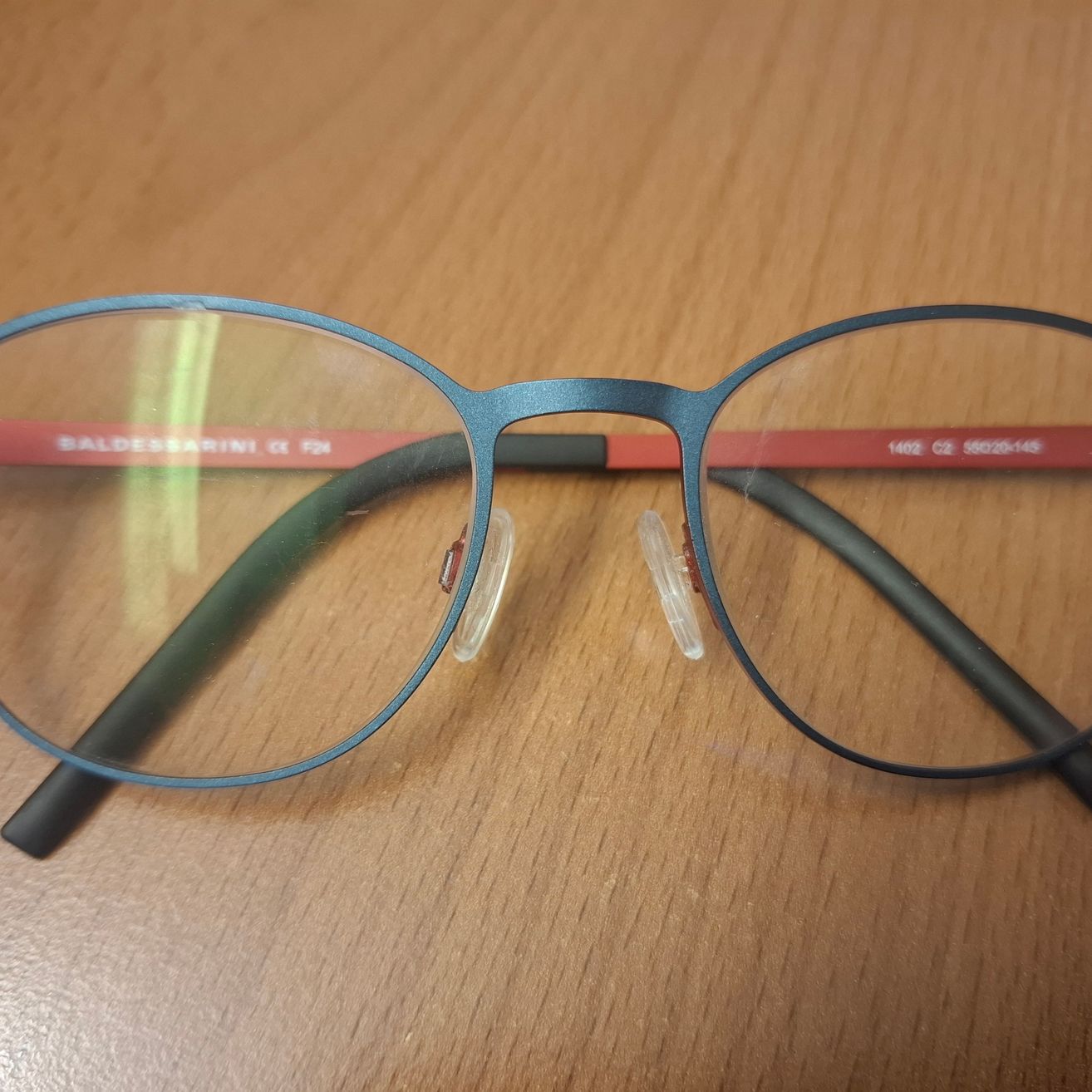 Ein Brillengestell mit blauen Bügeln und roten Akzenten auf einer Holzoberfläche. Der Markenname Baldessarini ist in Rot auf den Bügeln aufgedruckt.