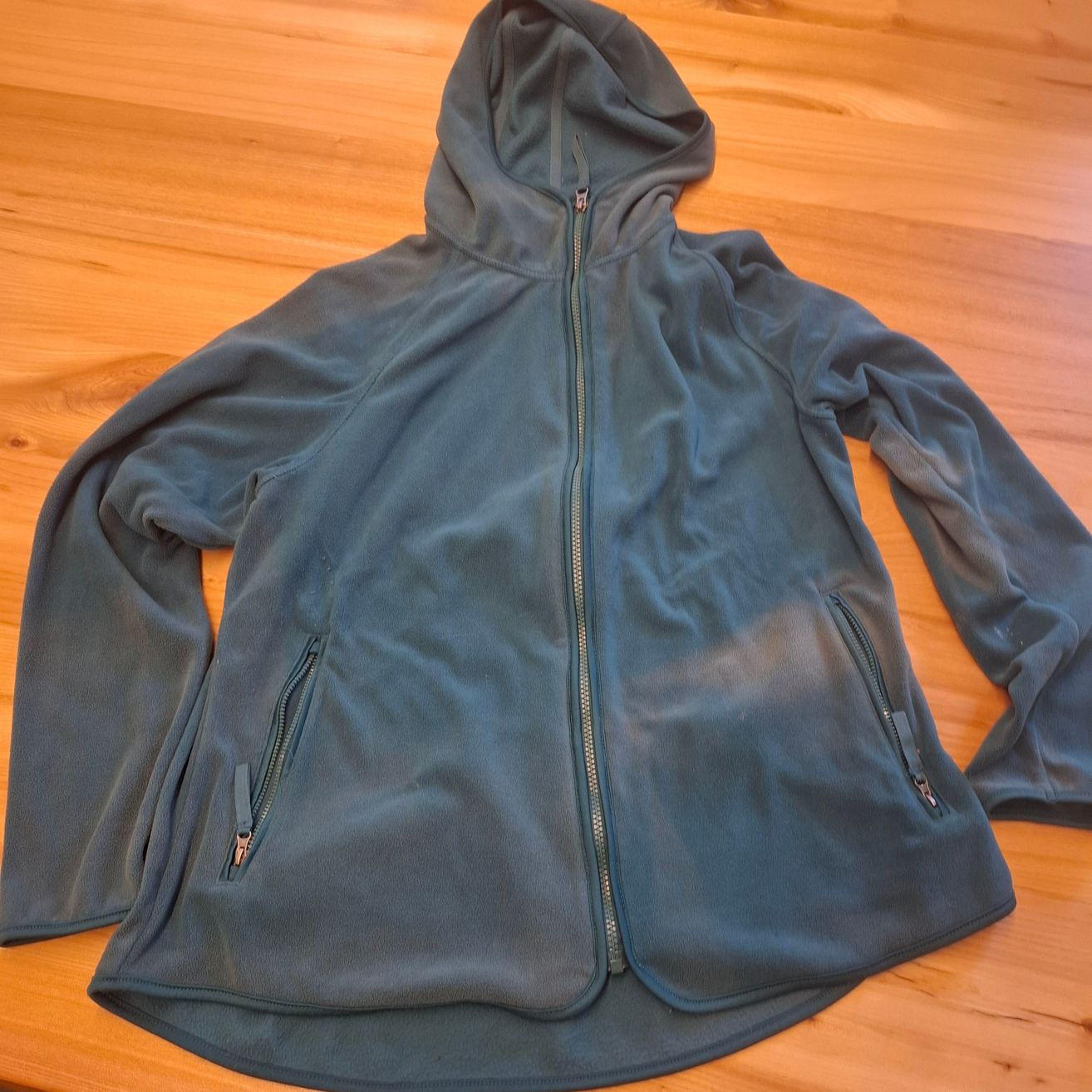 Eine hellblaue Fleecejacke mit Kapuze und Reißverschlüssen liegt flach auf einer Holzoberfläche. Die Jacke hat zwei Reißverschlusstaschen.