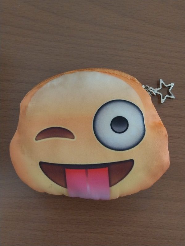 Nahaufnahme eines kleinen orangefarbenen Plüschtiers mit einem lächelnden Gesicht, großen Augen und einer herausgestreckten rosa Zunge auf einer Holzoberfläche.