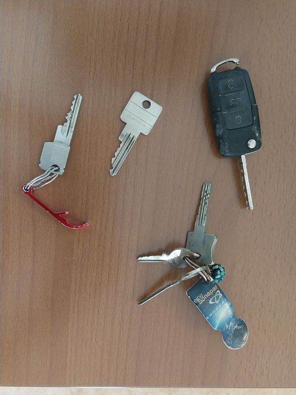 Fünf Schlüssel mit verschiedenen Schlüsselanhängern auf einer Holzoberfläche, darunter ein silberner Schlüssel, ein silberner Schlüssel mit rotem Clip, ein schwarzer Schlüsselanhänger, ein silberner Schlüssel mit blauem Schlüsselanhänger und ein silberner Schlüssel mit silbernem Schlüsselanhänger.