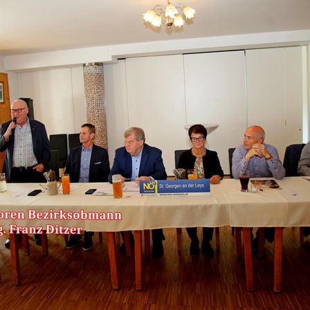 Eine Gruppe von Menschen sitzt bei einer Besprechung um einen Tisch. Eine Person spricht in ein Mikrofon. Auf dem Tisch stehen Gläser mit Getränken.