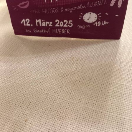 Eine lila Karte mit weißer Schrift auf einem Tisch. Es steht 'Madls Abend' und ist auf den 12. März 2025 datiert. Die Veranstaltung beginnt um 19:00 Uhr und findet im Gasthof Hueber statt.