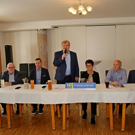 Eine Gruppe von Menschen sitzt um einen Tisch herum mit einem Mikrofon, einem Schild und Getränken. Ein Mann steht und spricht in das Mikrofon.