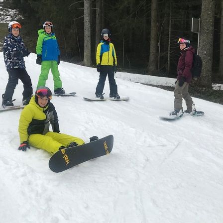 Vier Snowboarder stehen auf einem verschneiten Hang, einer in einem gelben Mantel sitzt, während die anderen mit Helmen und Handschuhen stehen.