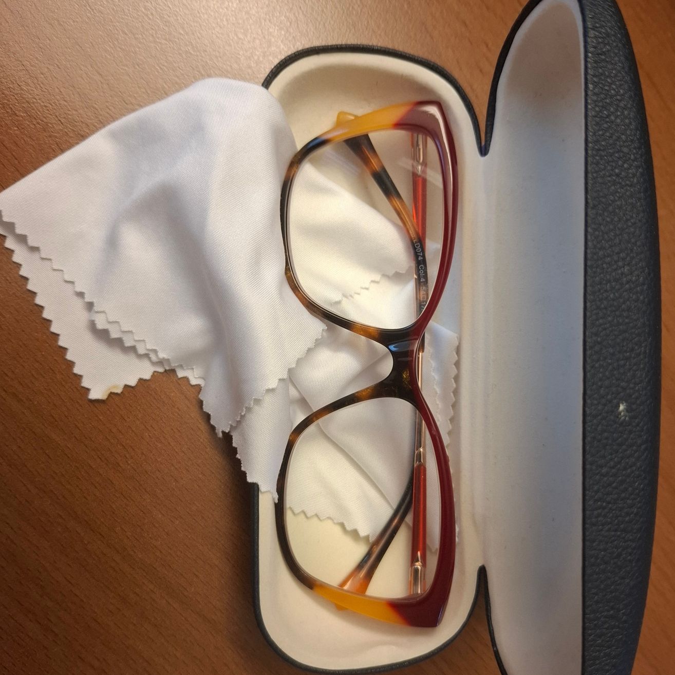 Ein Brillengestell mit roten Bügelenden befindet sich in einem geöffneten Etui mit einem weißen Tuch auf einem Holztisch.