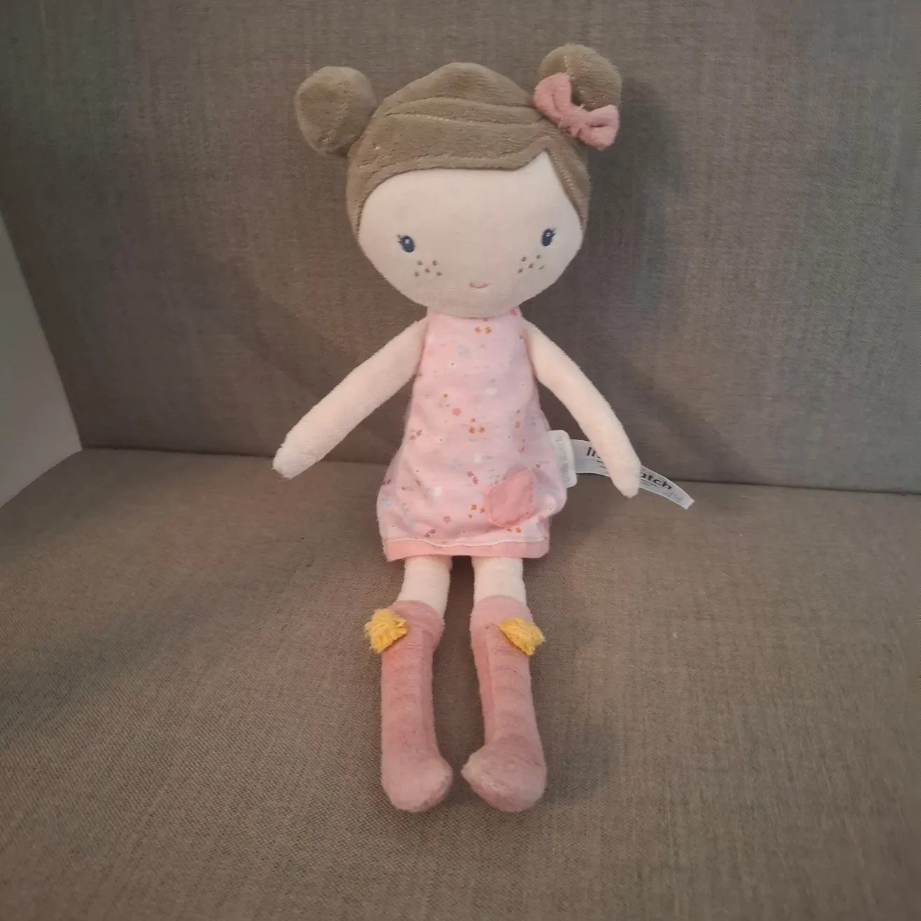 Eine weiche Spielzeugpuppe mit braunen Haaren, die ein rosa Kleid und rosa Stiefel mit gelben Etiketten trägt, sitzt auf einer grauen Couch.