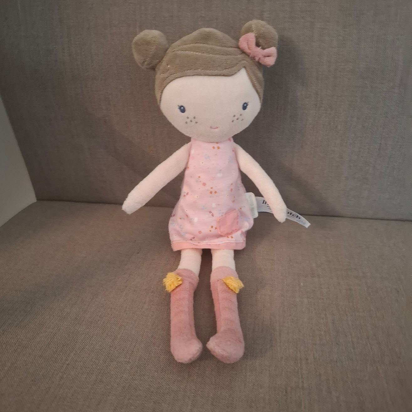 Eine weiche Spielzeugpuppe mit braunen Haaren, die ein rosa Kleid und rosa Stiefel mit gelben Etiketten trägt, sitzt auf einer grauen Couch.