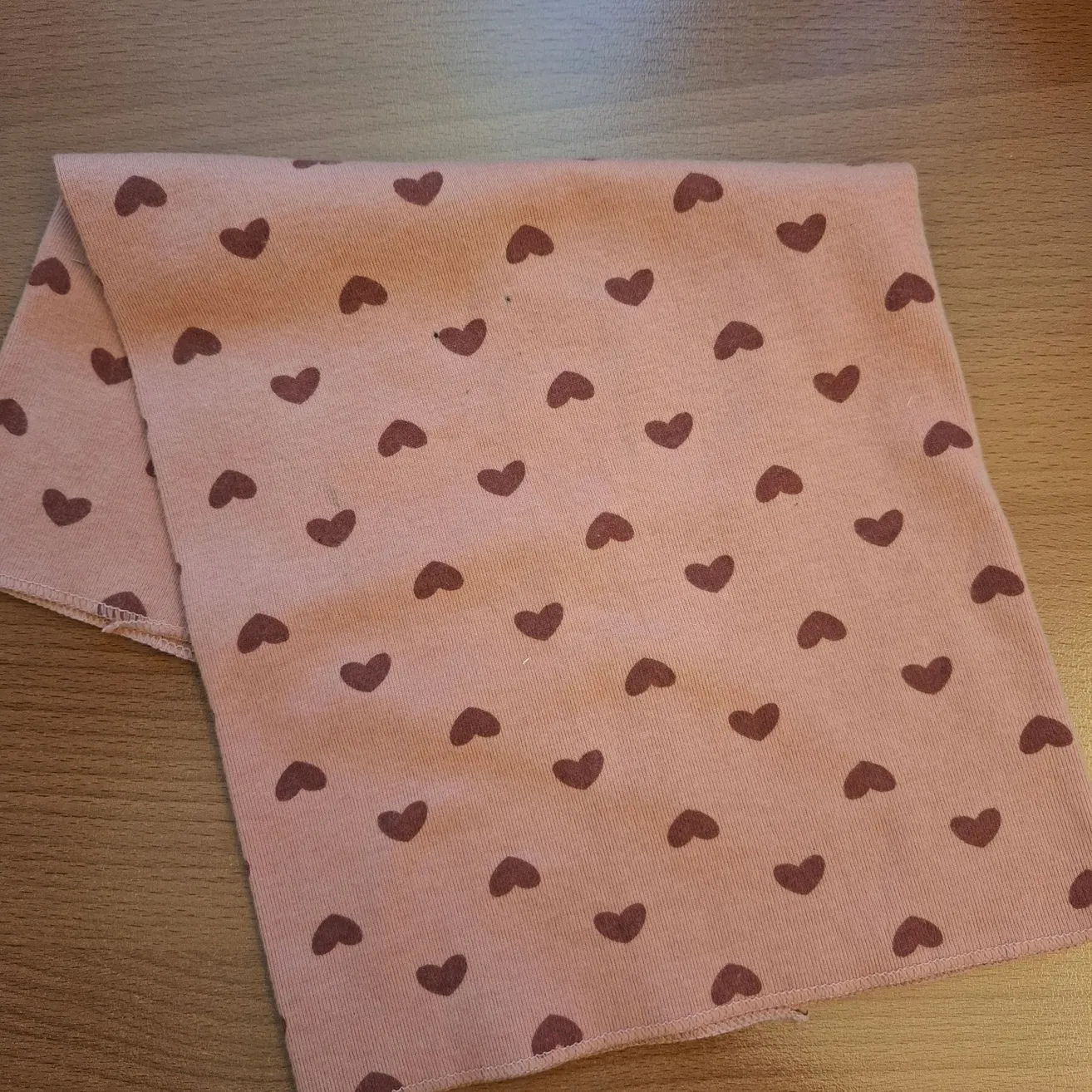 Ein rosa Tuch mit Herzensmuster, auf einer Holzoberfläche platziert. Das Herzensmuster ist braun, und der Stoff hat eine leicht strukturierte Oberfläche.