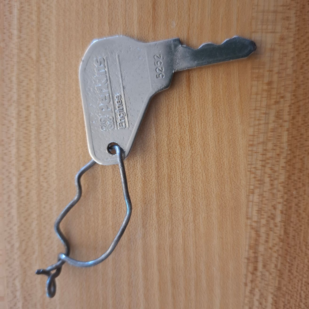 Ein silberner Schlüssel mit einem gebogenen Metallhaken, der an ihm befestigt ist und die Nummer 5252 trägt. Er liegt auf einer Holzoberfläche.