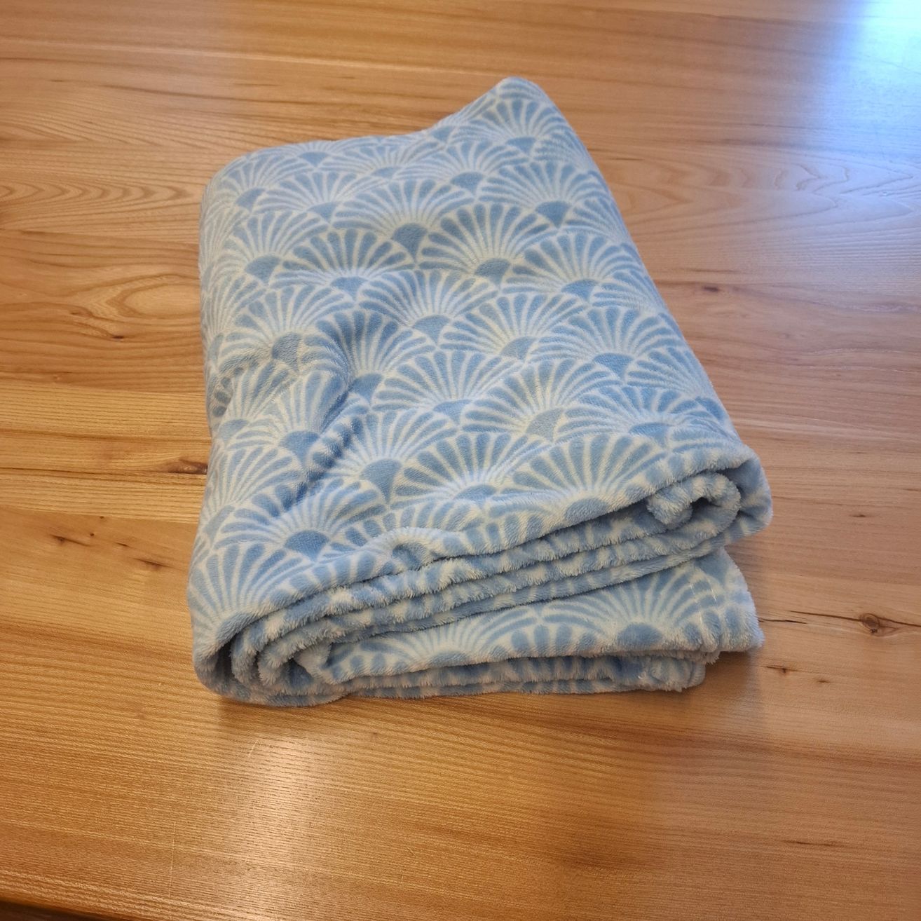Eine blaue Decke mit Muschelmuster ist gefaltet und auf einer Holzoberfläche abgelegt. Die Decke ist weich und warm, ideal für gemütliche Momente.