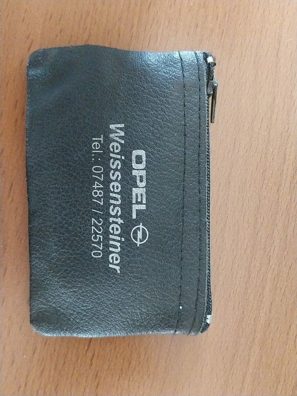 Ein schwarzes Lederportemonnaie mit der Marke Opel auf einer Holzoberfläche. Das Portemonnaie hat einen Reißverschlussverschluss und die Texte "Weissensteiner" und eine Telefonnummer "Tel: 07487/22570" sind darauf gedruckt.