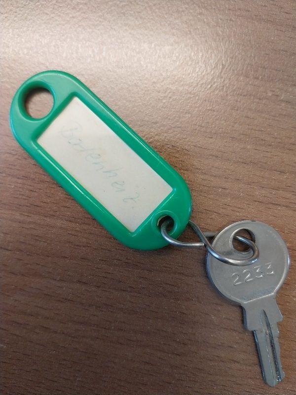 Ein grüner Schlüsselanhänger mit einem weißen Etikett auf einer braunen Holzoberfläche. Das Etikett zeigt den Text 'September 22' und der Schlüsselanhänger ist an einen Schlüssel mit der Bezeichnung '2233' befestigt.