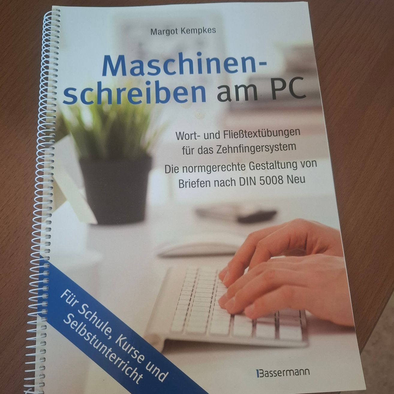 Ein Buch mit dem Titel 'Maschinen-schreiben am PC' ist auf einer Holzoberfläche zu sehen. Das Buchcover zeigt eine Hand, die auf einer Tastatur tippt, und einen Topf mit Pflanzen im Hintergrund.