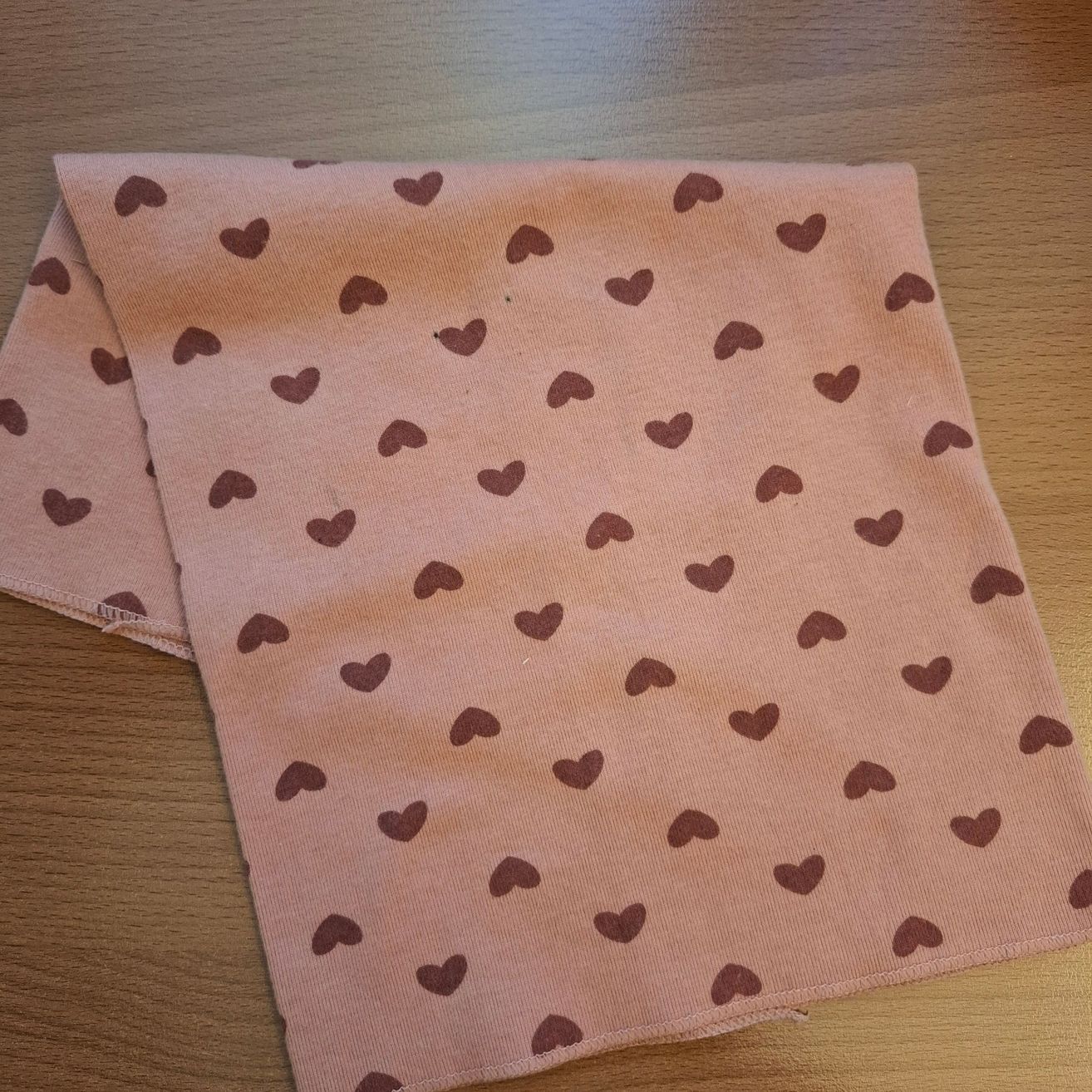Ein rosa Tuch mit Herzensmuster, auf einer Holzoberfläche platziert. Das Herzensmuster ist braun, und der Stoff hat eine leicht strukturierte Oberfläche.