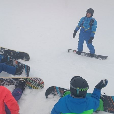 Eine Gruppe von Snowboardern, einige sitzend und andere stehend, sind auf einem schneebedeckten Hang.