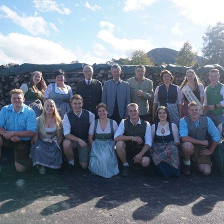Eine Gruppe von Menschen in traditioneller deutscher Kleidung posiert für ein Foto vor einer Steinmauer und einem Holzstapel.