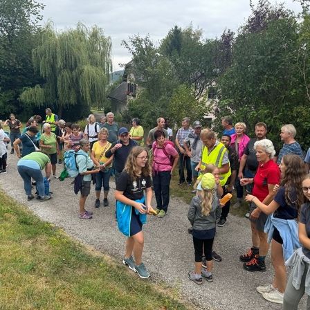 Eine Gruppe von Menschen steht auf einer Straße, vielleicht für eine Wanderung. Einige tragen Brillen und Turnschuhe, und andere haben Rucksäcke dabei. Eine Person hält ein gelbes Objekt. Ein goldenes Kreuz steht rechts.