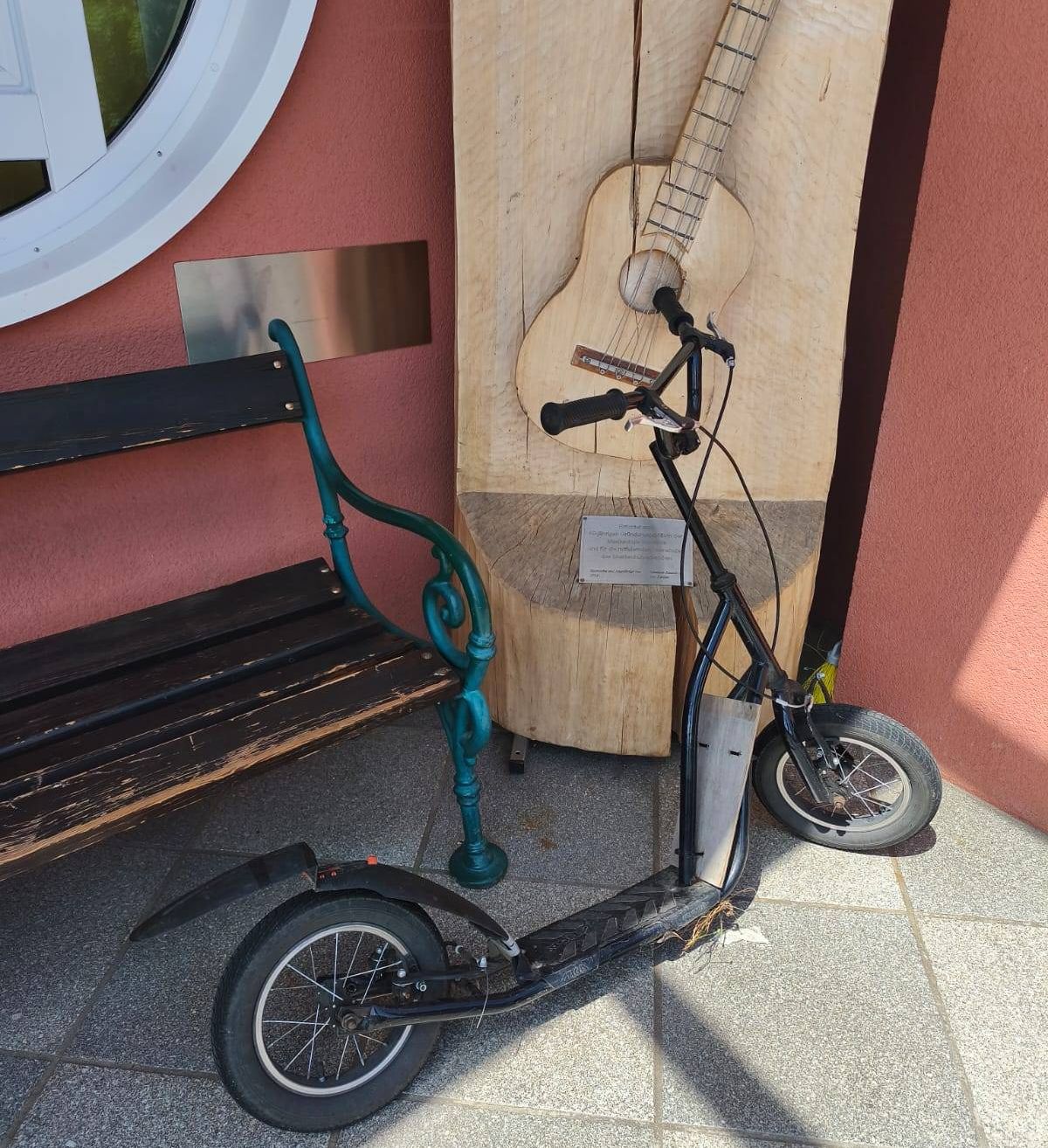 Ein schwarzer Elektroroller steht neben einer hölzernen Bank und einer großen Gitarrenskulptur an einer rosa Wand.