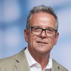 Ein Mann mit grauem Haar und einem Schnurrbart trägt eine Brille und eine helle Anzugjacke. Er steht vor einem blauen Himmel mit einem Logo auf seiner Jacke.
