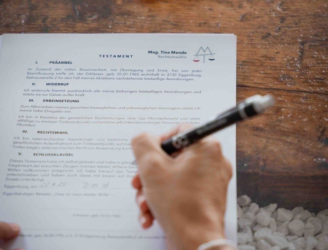 Eine Hand, die einen Stift hält, schreibt auf ein Dokument mit dem Titel 'Testament', das juristische Begriffe auf Deutsch enthält. Das Dokument liegt auf einem Holztisch mit Steinen in der Nähe. Der Text beinhaltet Details zu den letzten Wünschen einer Person und dem Datum des Schreibens.