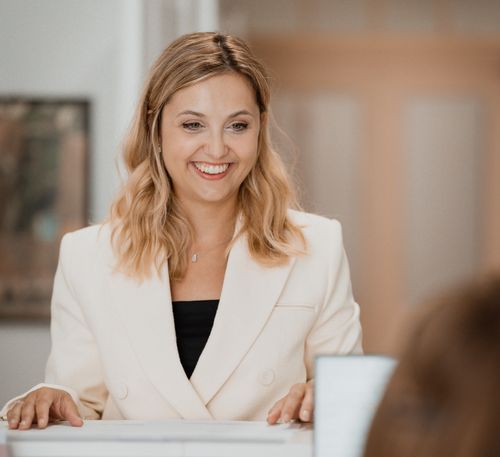 Eine lächelnde Frau in einem weißen Blazer und einem schwarzen Top sitzt an einem Schreibtisch, vielleicht in einem Büro. Sie schaut direkt in die Kamera.