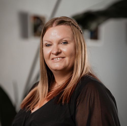 Eine lächelnde Frau mit blonden Haaren steht vor einer weißen Wand und trägt ein schwarzes Oberteil. Sie schaut direkt in die Kamera, vermutlich in einem Studio.