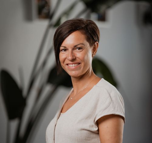 Eine Frau mit kurzen Haaren lächelt für ein Foto vor einer weißen Wand mit einer Pflanze. Sie trägt ein weißes Hemd und eine Halskette.