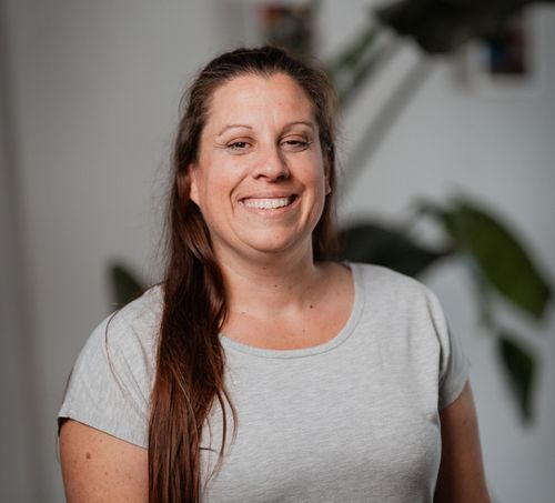 Eine lächelnde Frau mit langen braunen Haaren steht vor einer weißen Wand und einer grünen Pflanze. Sie trägt ein hellgraues T-Shirt.