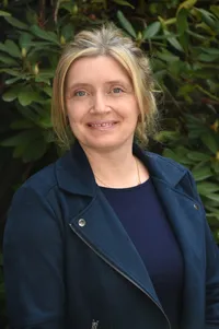 Eine Frau mit blondem Haar und einem Nasenring, die eine dunkelblaue Jacke und eine goldene Halskette trägt und für die Kamera lächelt.