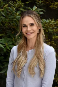 Eine Frau mit langen blonden Haaren, die für ein Porträt lächelt. Sie trägt einen hellblauen Cardigan, ein weißes Oberteil und eine herzförmige Halskette.