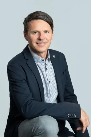 Ein Mann in einem dunkelblauen Blazer und hellblau kariertem Hemd mit einer Anstecknadel am Blazer lächelt in die Kamera.