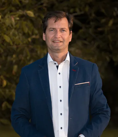 Ein Mann steht und lächelt, trägt einen marineblauen Blazer mit einem weißen Hemd und einem roten Etikett in der linken Tasche. Er befindet sich in einer Außenumgebung mit einem unscharfen Hintergrund von Bäumen.