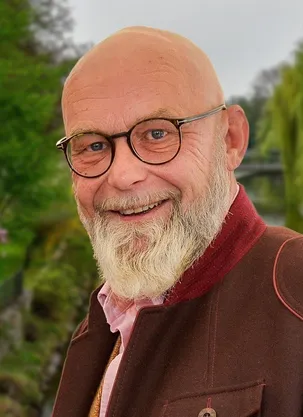 Ein älterer Mann mit weißem Bart und Brille lächelt. Er trägt ein rosa Hemd unter einer braunen Jacke. Im Hintergrund befinden sich Bäume und Grünpflanzen.