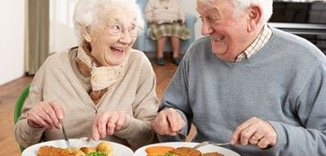 Zwei ältere Menschen sitzen an einem Tisch, lächeln und genießen ihr Essen. Sie haben jeweils einen Teller mit Essen vor sich. Die Frau trägt eine Brille und einen Schal, der Mann einen grauen Pullover. Im Hintergrund sitzt eine weitere Person auf einem Stuhl.
