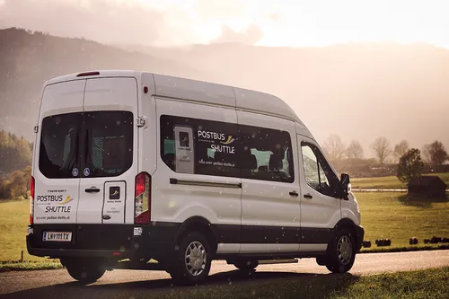 Ein weißer Postbus Shuttle Van fährt auf einer Straße mit grasbewachsenen Feldern und Bergen im Hintergrund.