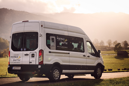 Ein weißer Postbus Shuttle Van fährt auf einer Straße mit grasbewachsenen Feldern und Bergen im Hintergrund.