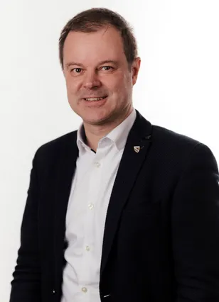 Ein lächelnder Mann in einem weißen Hemd und einer schwarzen Jacke mit einer Anstecknadel am linken Revers posiert für ein Foto vor weißem Hintergrund.