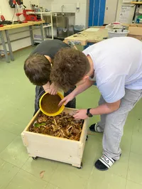 Zwei Jungen arbeiten zusammen, einer gießt Erde in einen gelben Eimer, während der andere einen Behälter mit einer Mischung hält. Sie sind in einem Raum mit einem Holzwagen, der mit Blättern und Erde gefüllt ist.