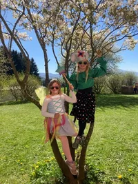 Zwei Mädchen, eines als Fee verkleidet, sitzen auf einem Ast unter einem blühenden Baum und posieren für ein Foto an einem sonnigen Tag.