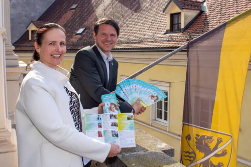 Zwei Menschen lächeln und halten Broschüren vor einem Gebäude. Eine ist eine Frau in einem weißen Mantel, der andere ein Mann in einem Anzug. Sie stehen auf einer Steinkante.