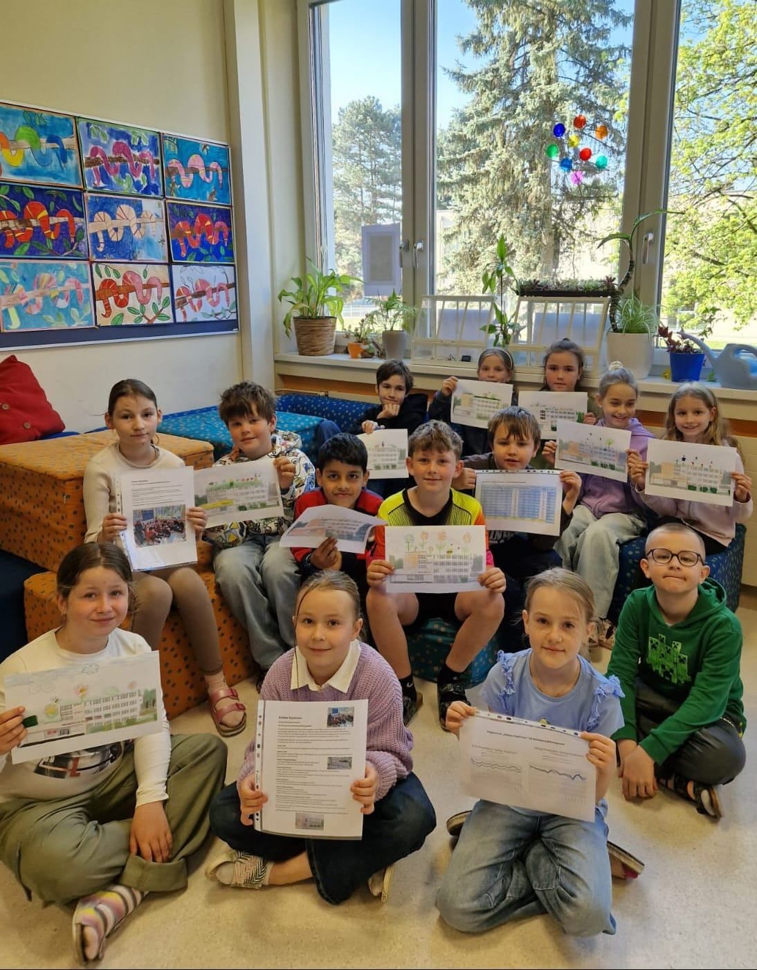 Eine Gruppe von Kindern sitzt auf dem Boden, hält Papiere hoch und lächelt in einem Klassenzimmer mit Blick auf Bäume vor dem Fenster.