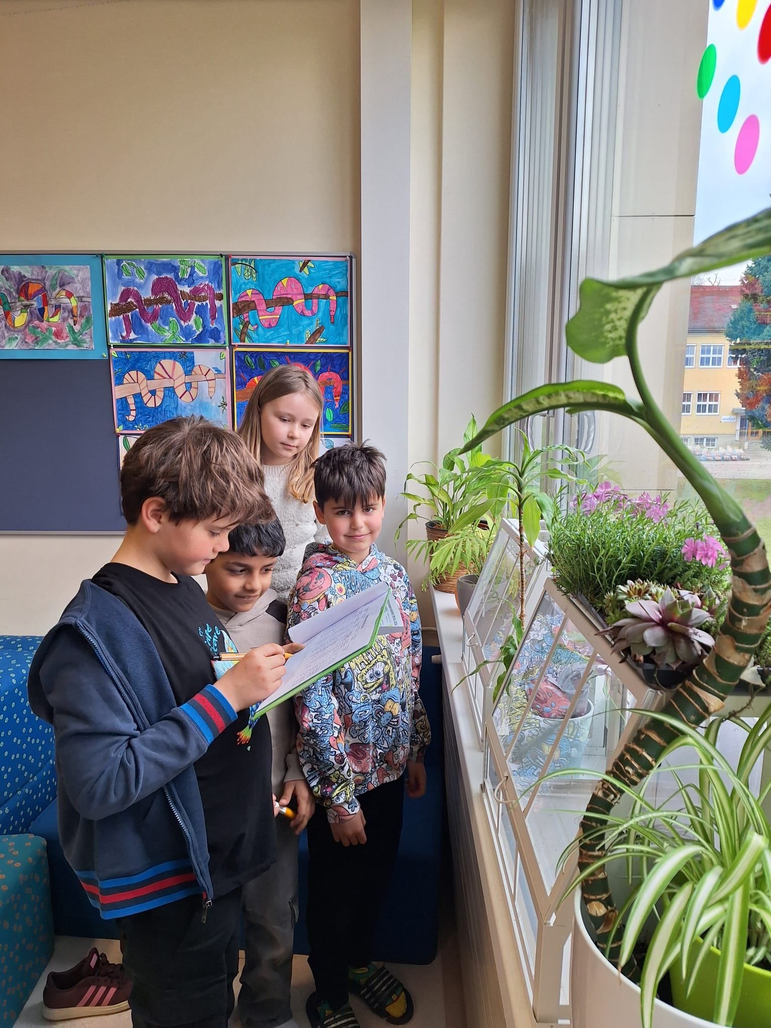 Vier Kinder stehen in einem Klassenzimmer, wobei ein Junge ein Stück Papier in der Hand hält und die anderen einen Pflanzenkasten betrachten. Dahinter hängen bunte Gemälde an der Wand.