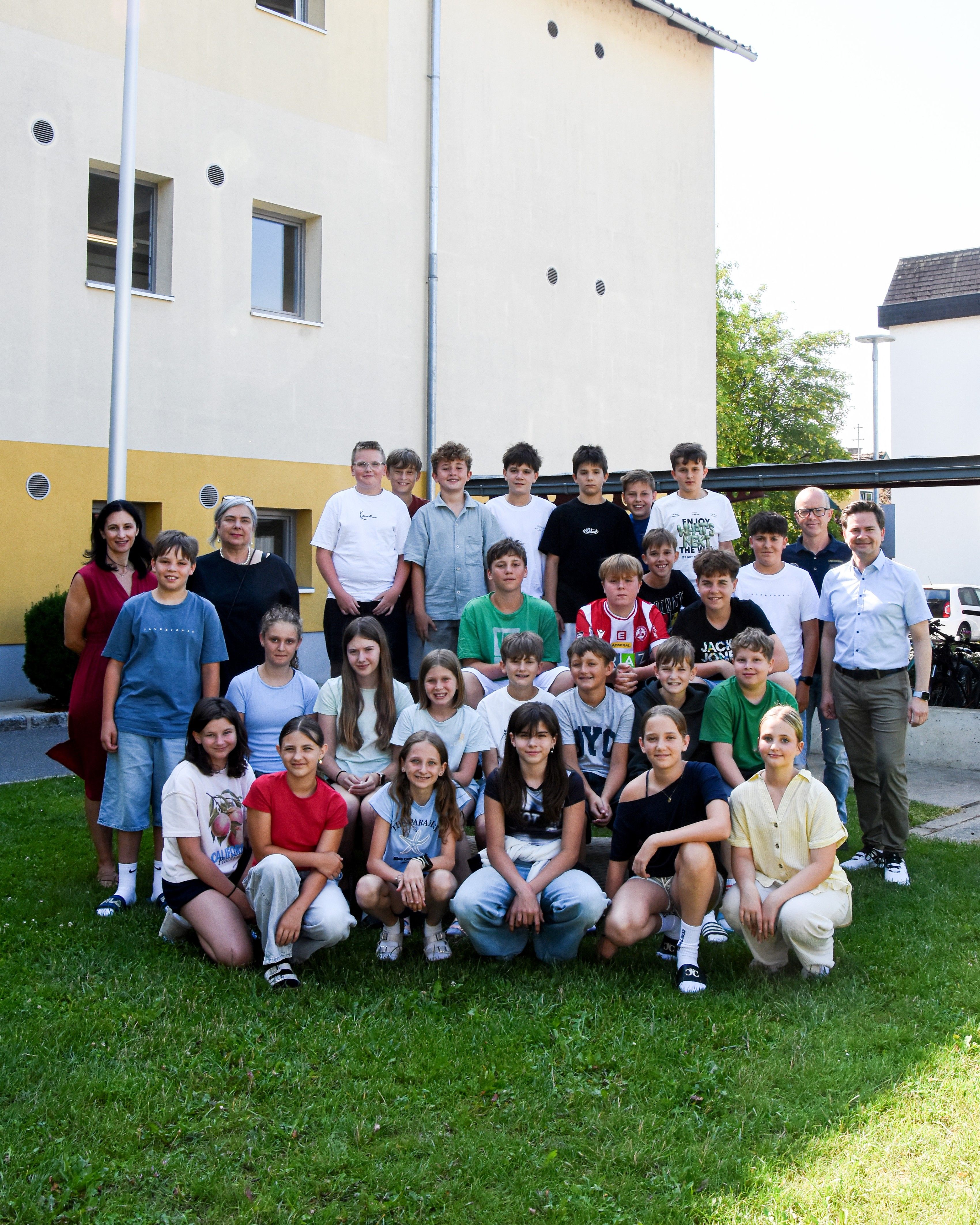 Eine Gruppe von Kindern und Erwachsenen posiert für ein Foto auf dem Rasen vor einem Gebäude. Sie tragen alle verschiedenfarbige Kleidung.