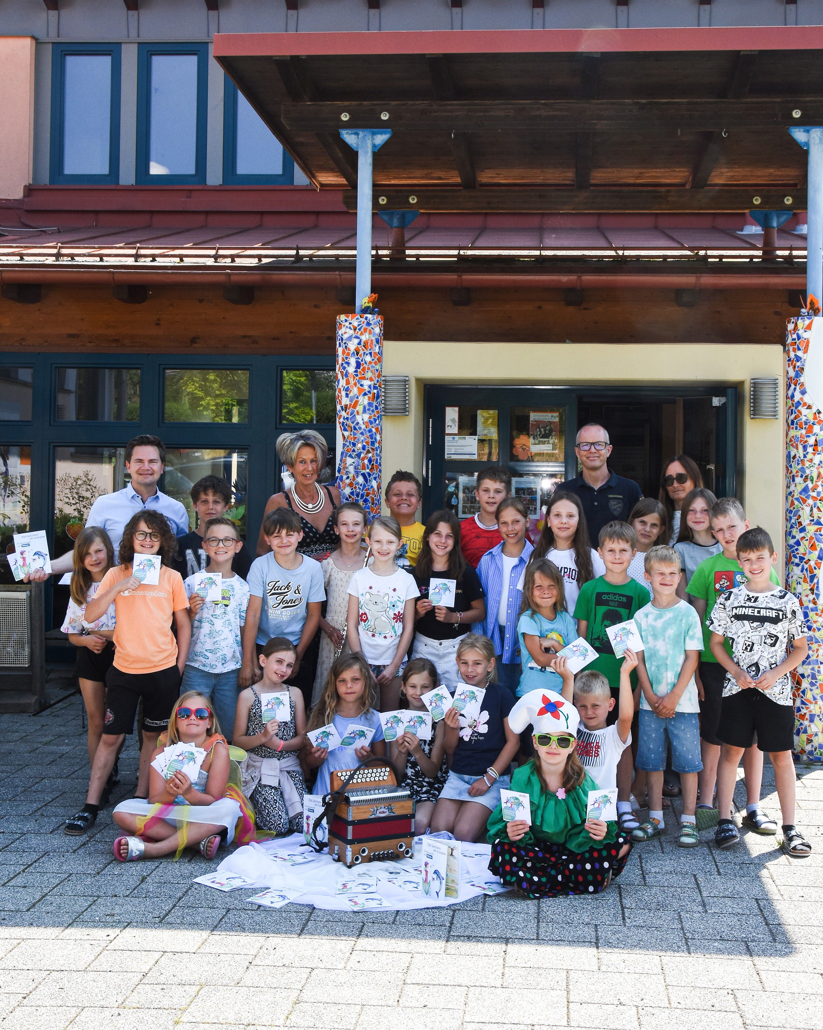 Eine Gruppe von Kindern und Erwachsenen versammelt sich vor einem Gebäude. Sie halten Karten in der Hand und lächeln für die Kamera. Zwei Personen sitzen auf dem Boden.