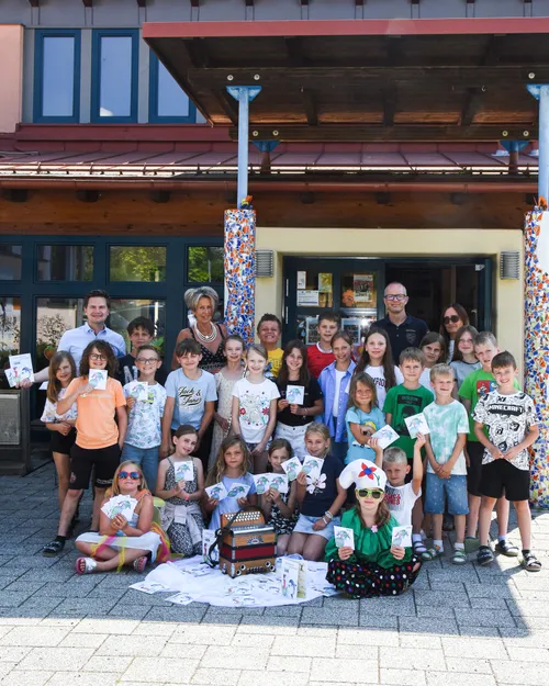 Eine Gruppe von Kindern und Erwachsenen versammelt sich vor einem Gebäude. Sie halten Karten in der Hand und lächeln für die Kamera. Zwei Personen sitzen auf dem Boden.