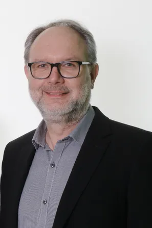 Ein Mann mit Bart und Schnurrbart trägt eine Brille und einen schwarzen Anzug. Er hat ein graues kariertes Hemd darunter. Er lächelt und scheint für ein Foto zu posieren.